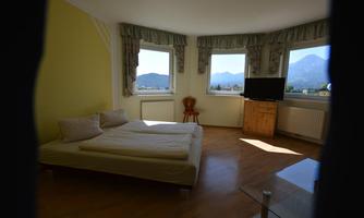 A cozy bedroom with large windows and a view of the mountains. There is a bed, a television, and a wooden chair.