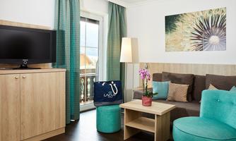 A cozy living environment with gray and turquoise furniture. The room features a television, a balcony, and large windows with curtains.