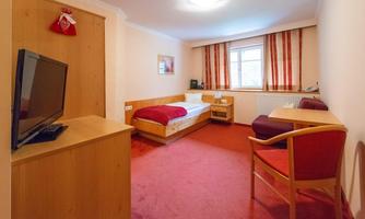 A simple hotel room with a single bed and a small table. The room has a television and a friendly red carpet.