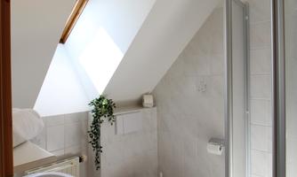 A modern bathroom with a slanted ceiling and plenty of natural light. It is bright and clean, equipped with a shower, toilet, and sink.