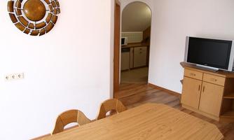 A cozy living area with a wooden table and several chairs. In the background, there is a television and a kitchen corner.