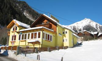 A yellow guest house in a snow-covered landscape. In the background, mountains and a blue sky can be seen.