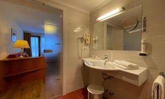 A modern bathroom with a sink, mirror, and shower. A stool is placed next to the sink, and in the background, a bright room with a table is visible.