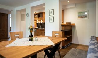 A cozy dining area with a wooden table and chairs. On the table, there is a small flower vase and a decorative cloth.