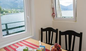 A cozy dining table with fresh fruits and decorations stands in front of a window overlooking the lake. The warm colors and pleasant light create an inviting atmosphere.