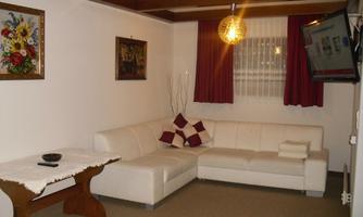 A cozy living room with a white sofa and a wooden table. Pictures hang on the walls, and the window is adorned with red curtains.