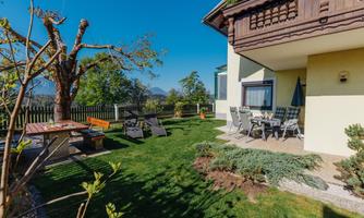 A cozy garden with lawn, a table and chairs. In the background, there is a house with a balcony and blue sky.
