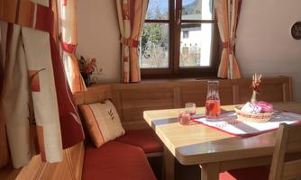 A cozy dining area with a wooden bench and red fabric. A table with decoration, drink, and a view out of the window onto a bright, friendly environment.