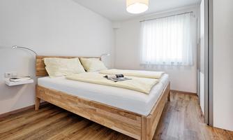 A simple, modern bedroom with a wooden bed and two pillows. The room is bright and friendly, with a large window and a warm wooden floor.