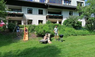 A large residential building with a well-maintained garden. Some people are enjoying the day outdoors while a grill is set up.