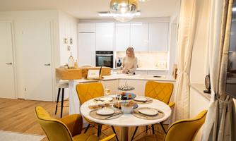 A modern kitchen with a round dining table and elegant yellow chairs. In the background, there is a woman working at the cooking area.