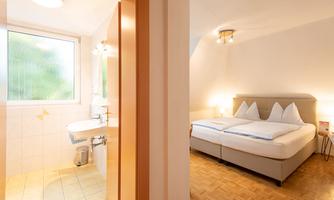 A bright bedroom with two beds and a window. Next door is a clean bathroom with a sink and shower.