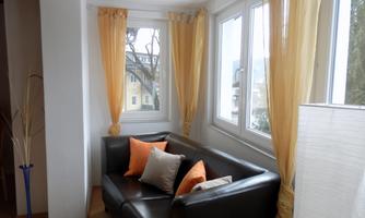A cozy seating area with a black sofa and colorful cushions. Large windows let in plenty of light, and the yellow curtains create a friendly atmosphere.