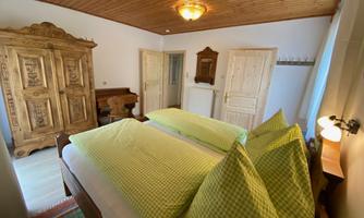 A cozy bedroom with two beds and green cushions. In the background, there is an antique wardrobe and the walls are bright and friendly.
