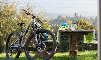 A mountain bike stands next to a table outdoors. On the table are a helmet and glasses, surrounded by green nature.