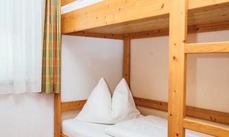 A simple room with a wooden bunk bed. The bedding is clean and tidy, and a window lets in daylight.