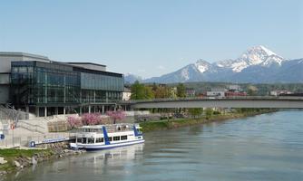 A modern building view by the river with a boat in the foreground. In the background, snow-capped mountains can be seen.