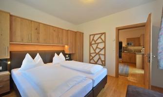 A modern bedroom with two single beds and wooden wardrobes. In the background, an open kitchenette can be seen.