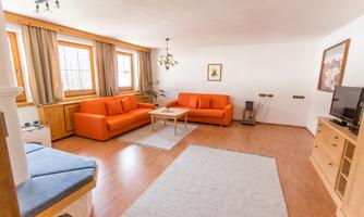 A bright living room with an orange sofa set and large windows. A wooden table is in the center, next to it a television and a sideboard.