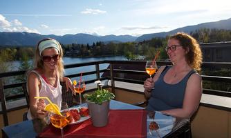 Two women are enjoying cocktails on a balcony overlooking a lake and the mountains. The table is decorated with fresh strawberries and a small plant.