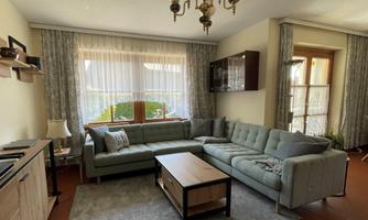 A bright living room with a comfortable gray sofa and a table in the center. Large windows provide plenty of natural light.