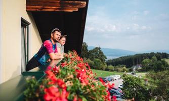 A man and a boy are standing on a balcony, looking at the landscape. In the foreground, red flowers are blooming, while mountains and trees can be seen in the background.