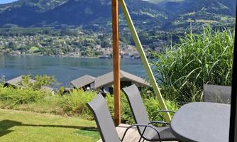 A terrace with modern chairs and a table overlooks a picturesque lake. The sky is blue with some clouds and the surroundings are green.