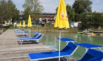 A relaxed waterfront promenade with blue loungers and yellow beach umbrellas. In the background, the water and some trees can be seen.