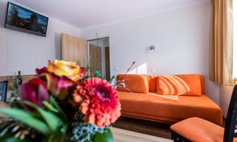 A cozy room with an orange sofa and a TV on the wall. A floral arrangement in the foreground adds color.