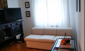 A bright living room with a white sofa and a black TV cabinet. The windows are covered with white curtains and the floor is made of wood.