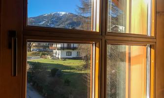 A window view of a picturesque landscape with mountains and a clear blue sky. Outside, green meadows and a few houses are visible.