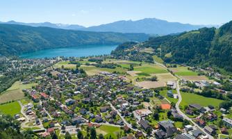 A picturesque village at the foot of the mountains, surrounded by green fields and a clear lake. The surroundings are peaceful nature and invite relaxation.