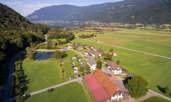 An idyllic rural landscape with expansive meadows and a small village. In the background, mountains and a lake are visible.