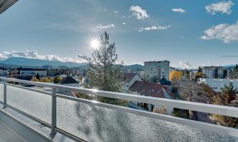 A beautiful view from a balcony with sunlight and clouds. In the background, buildings and mountains are visible.