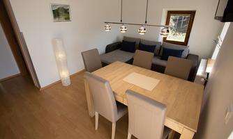 A modern dining room with a wooden table and beige chairs. In the background, there is a comfortable sofa and a window that lets in daylight.