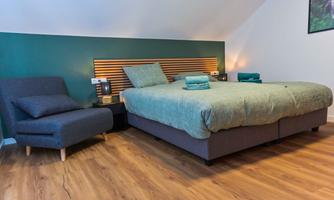 A modern bedroom with a comfortable bed and a cozy armchair. The walls are painted in soft green tones, giving the room a relaxed atmosphere.