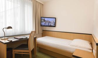 A simple hotel room with a single bed, a desk, and a television mounted on the wall. The room has large windows that let in plenty of daylight.