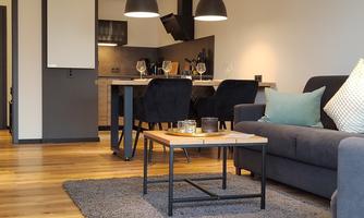A modern living room with a gray sofa and a wooden table. In the background, an open kitchen with stylish lighting and a cozy atmosphere is visible.