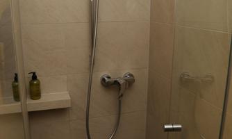A modern shower with glass walls and a wall-mounted hand shower. The walls are in neutral colors, and there are some hygiene products on a small shelf.