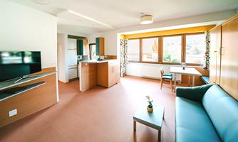 A modern, bright living room with a kitchen and large window. There is a comfortable sofa, a table, and a television.