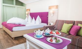 A cozy room with a large bed and a colorful sofa. On a table, there is a teapot and some small snacks.