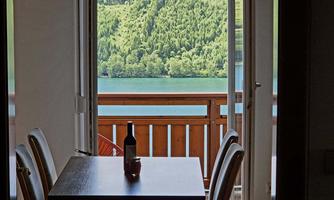 A beautiful view of a clear lake and wooded hills from a balcony. A wine bottle is on the table.