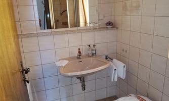 A small, clean bathroom with a sink and a mirror. On the floor, there is a stool and two towels are hanging within reach.