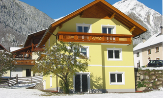 A yellow house with a balcony in a snowy landscape. In the background, you can see mountains and a clear blue sky.