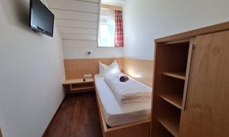 A simple room with a single bed and a television on the wall. The room has light wood and a window with curtains.