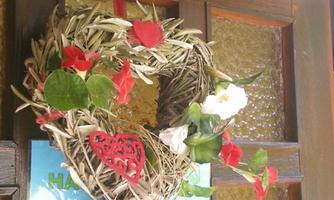 A heart-shaped wreath made of plants and flowers hangs on a door. It is adorned with red and white blossoms, creating an inviting atmosphere.