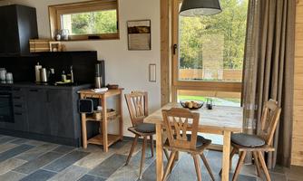 A modern kitchen with a dining area made of wood. Large windows provide a view of the outdoors.