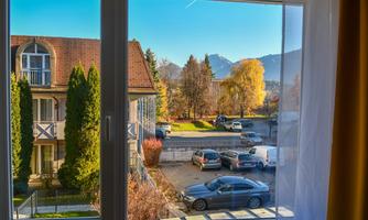 A view from a window onto a scenic landscape with mountains in the background. Parked cars and colorful deciduous trees are visible.