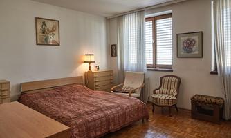 A bedroom with a large bed, a nightstand, and a chair. There are windows with curtains and paintings on the walls.