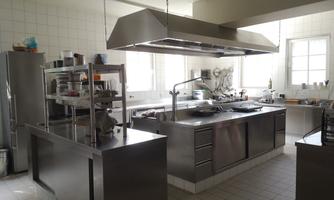 A modern kitchen with stainless steel surfaces and large windows. The equipment includes a grill and shelves for kitchen utensils.